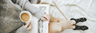 woman holding a cup of coffee at right hand and reading book on her lap while holding it open with her left hand in a well-lit room